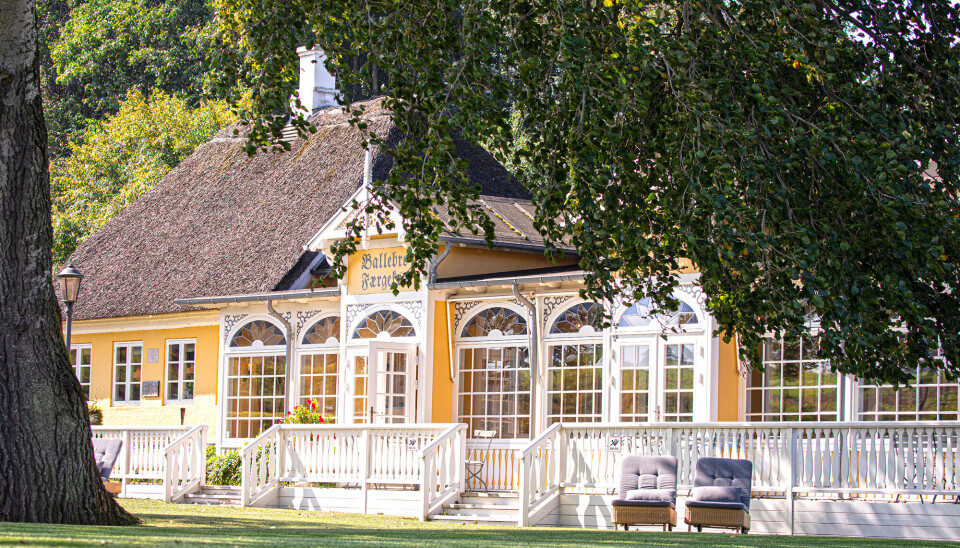 Et foto af Ballebo Kro set ude fra. Det er en gul bygning, med stråtag. Der er mange, store og høje vinduer, og to stole nedenfor terrassen.
